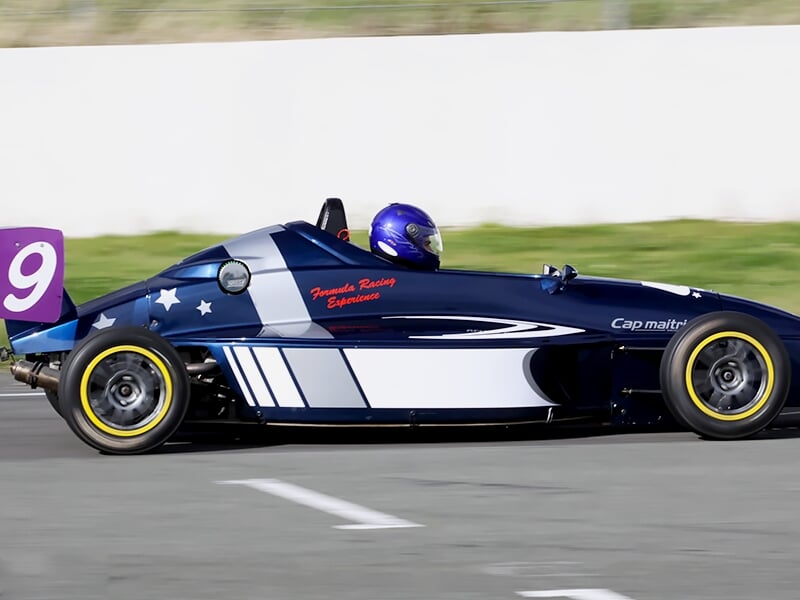 Voiture de course monoplace bleue et blanche sur circuit, vue latérale avec un pilote en combinaison bleue.