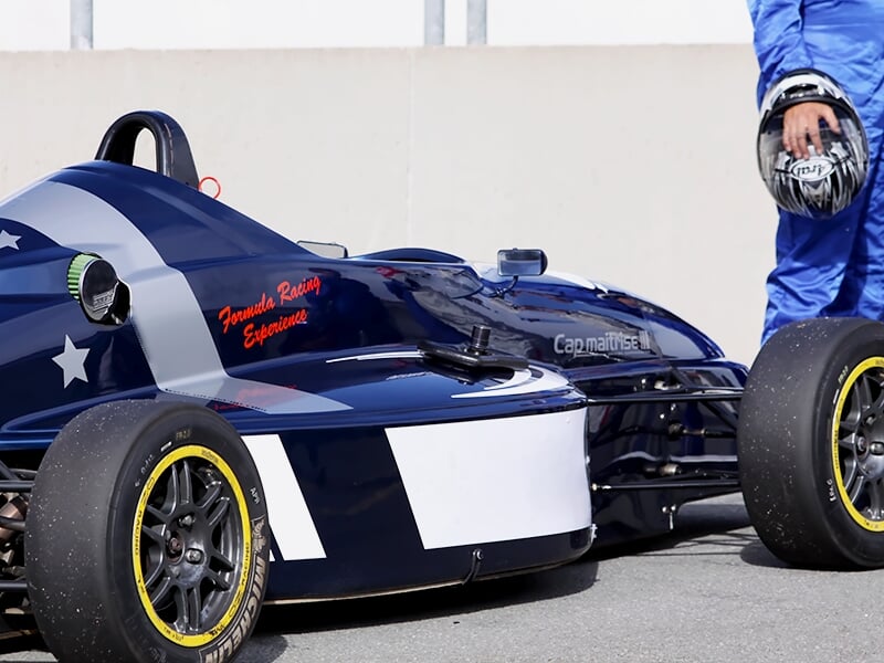 Voiture de course monoplace bleue et blanche vue de près, avec un pilote debout à côté tenant un casque.