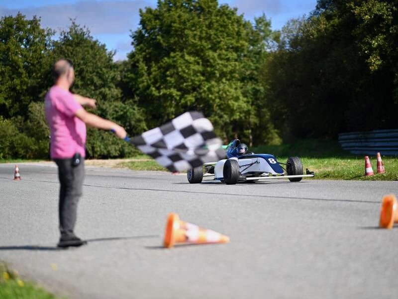 Voiture de course monoplace bleue sur circuit, drapeau à damier agité par une personne en rose.