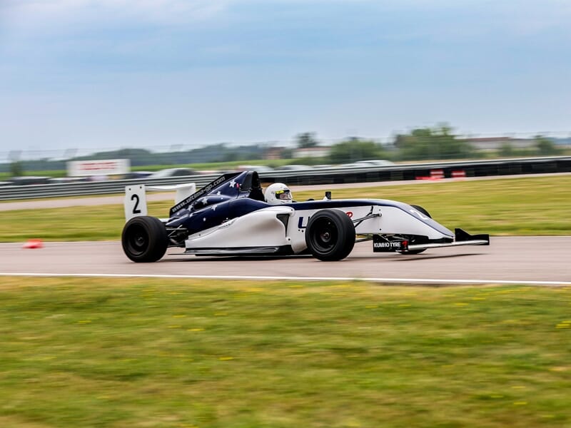 Voiture de course Formule 4 au circuit de Pont l'Evêque en pleine vitesse sur circuit avec herbe au premier plan.