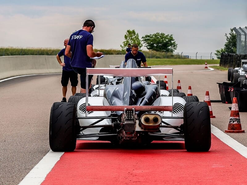 Vue arrière d'une Formule 4 au circuit de Pont l'Evêque avec trois personnes en tenue staff à proximité sur zone rouge.