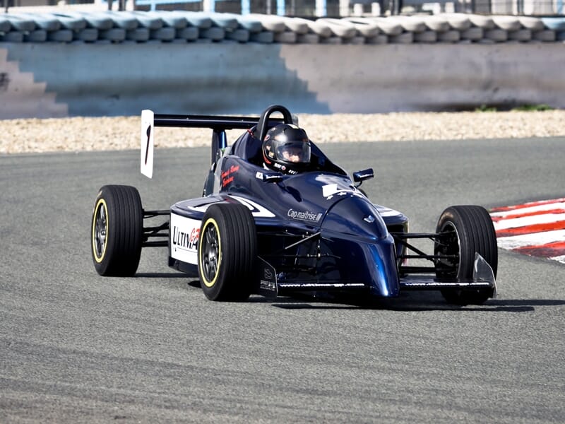 Formule Renault au circuit de Pont l'Evêque noire sur piste de course, pilote casqué en tenue noire, virage serré sur circuit asphalté.