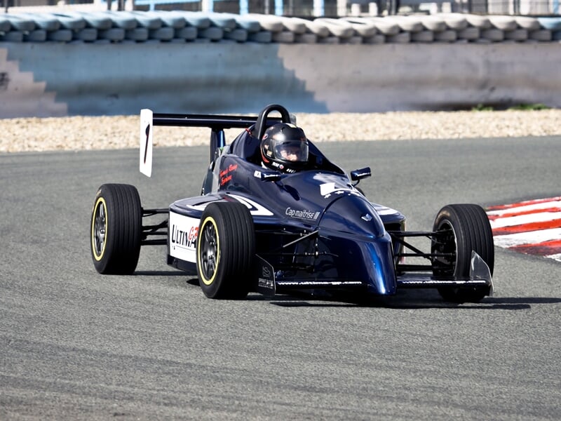 Formule Renault au circuit de Pont l'Evêque noire sur piste de course, pilote casqué en tenue noire, virage serré sur circuit asphalté.