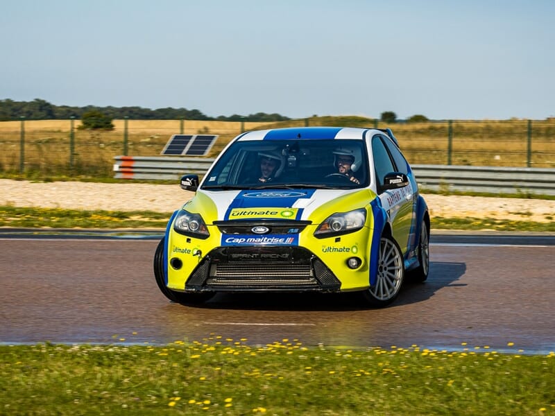 Voiture de course jaune et bleue sur circuit, prise de virage serré avec pilote concentré au volant.