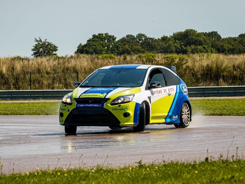 Voiture de rallye jaune et bleue sur piste mouillée, en virage avec paysage vert et barrières en arrière-plan.
