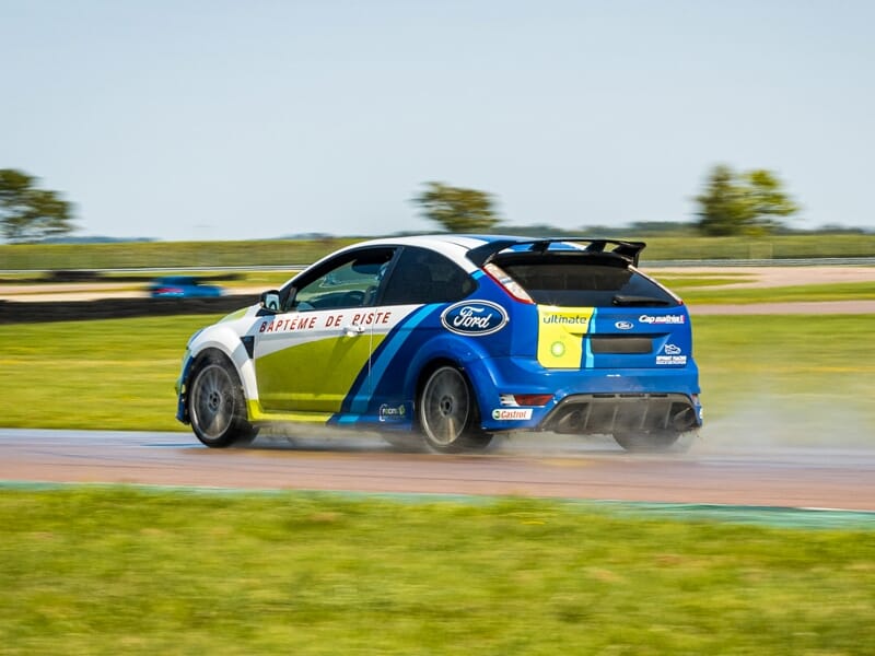 Voiture de course bleue, blanche et jaune dérapant sur circuit, avec fumée de pneus et herbe au premier plan.