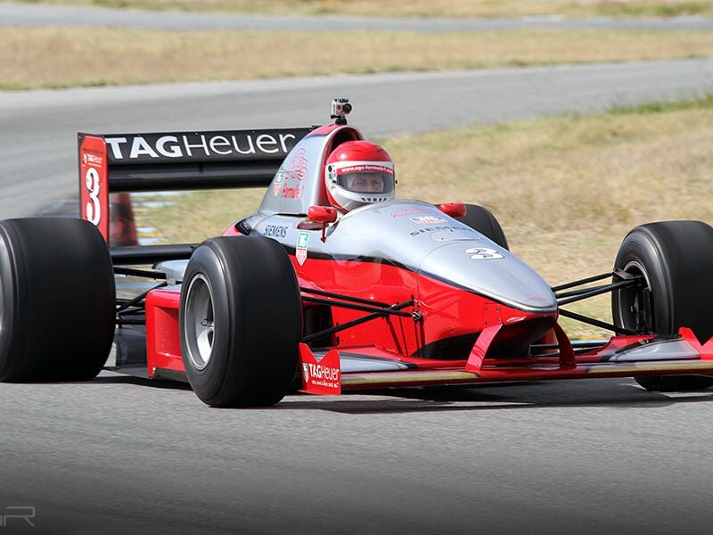 Voiture de course rouge et blanche de type monoplace sur circuit, en pleine vitesse avec un pilote casqué.