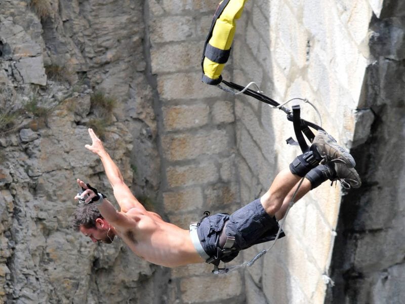 Homme torse nu effectuant un saut à l'élastique, en position tête en bas, depuis un pont en pierre surplombant des rochers