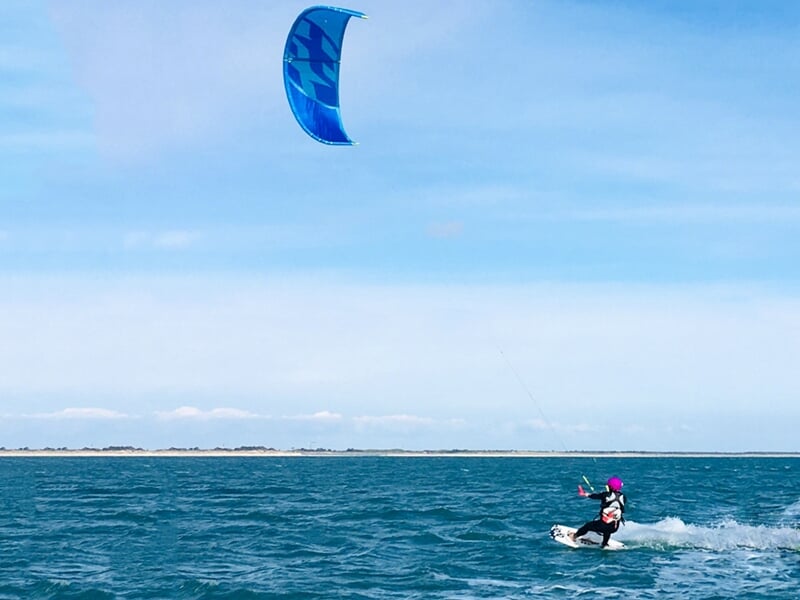 Personne pratiquant le kitesurf sur l'eau avec une voile bleue sous un ciel clair à l'Île de Ré.