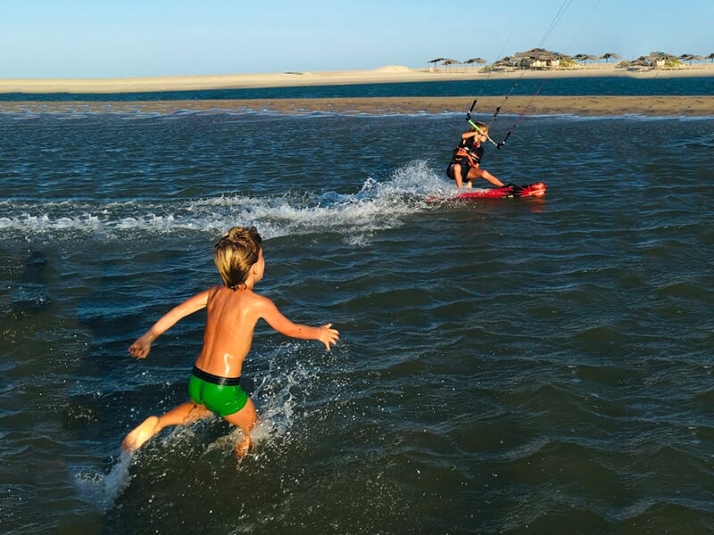 Enfant courant dans l'eau peu profonde vers un kitesurfeur en action près d'une plage sableuse.