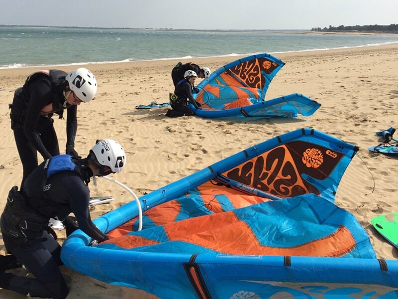 Trois personnes sur la plage préparant des voiles de kitesurf bleues et orange avant la session.
