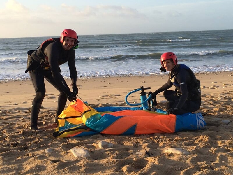 Deux kitesurfeurs souriants sur la plage avec leurs voiles colorées posées sur le sable.