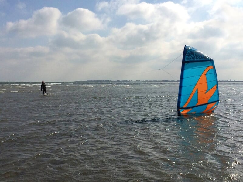Voile de kitesurf bleue et orange flottant dans l'eau peu profonde avec un kitesurfeur en arrière-plan.