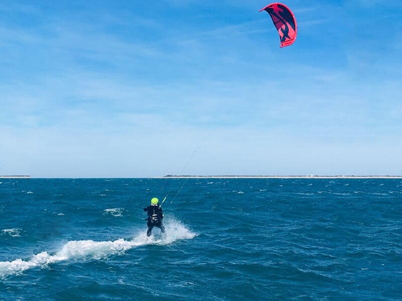Kitesurfeur en combinaison noire glissant sur l'eau avec une voile rouge et noire sous un ciel bleu.