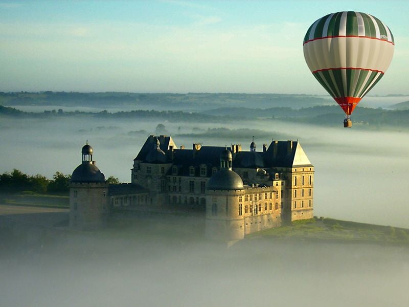 Montgolfière volant au-dessus d'un château entouré de brume matinale dans un paysage verdoyant.