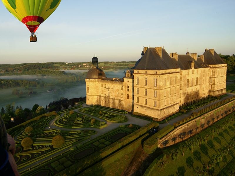 Montgolfière colorée survolant un grand château avec des jardins bien entretenus au lever du soleil.