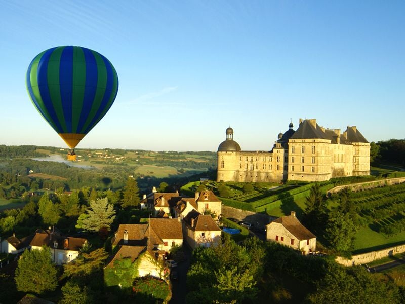 Montgolfière colorée survolant un village et un château dans un paysage vallonné et ensoleillé.