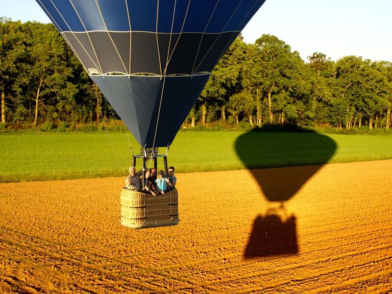 Montgolfière bleue avec passagers survolant un champ cultivé, ombre projetée au sol.