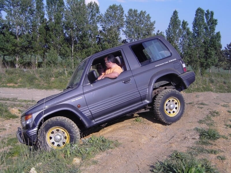 Homme conduisant un 4x4 gris sur un terrain accidenté avec herbe et terre.