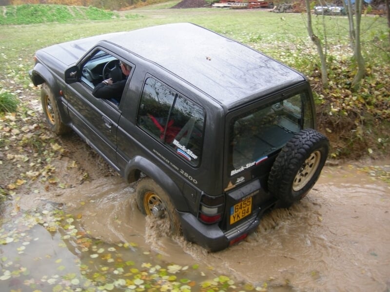 4x4 noir foncé roulant dans une flaque d'eau boueuse avec des feuilles flottantes.