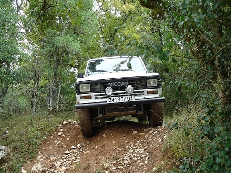Nissan 4x4 blanc franchissant un sentier escarpé en forêt, calandre tournée vers l’objectif, sol rocailleux et végétation dense.