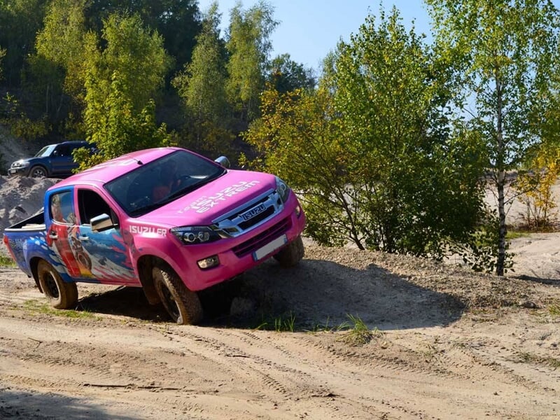 SUV rose et bleu évoluant sur un terrain sablonneux avec végétation et arbres.