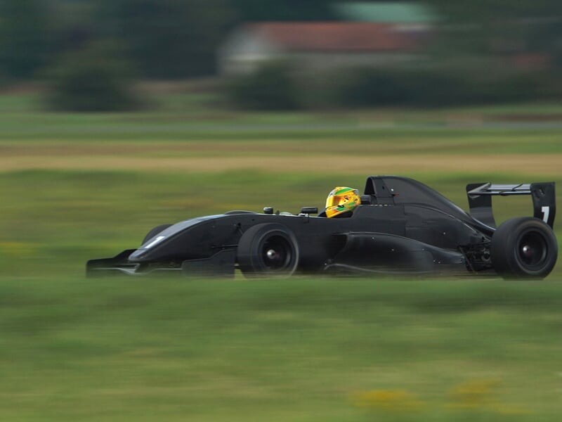 Voiture de course noire avec pilote en casque jaune roulant à grande vitesse sur circuit vert.