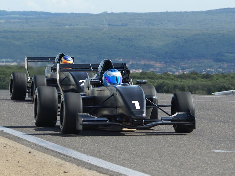 Deux monoplaces noires avec pilotes casqués roulant sur une piste de course en extérieur.