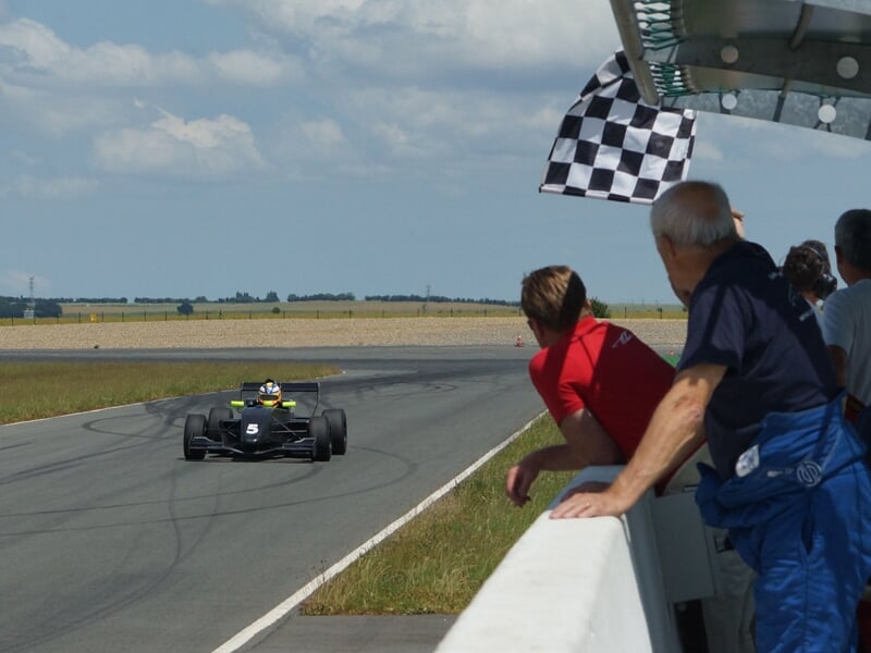 Spectateurs regardant une monoplace noire numéro 5 passer sous un drapeau à damier sur circuit.