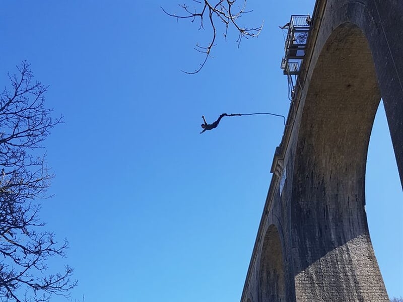 Une silhouette se balance au bout d'un élastique depuis un pont massif avec un ciel bleu clair en arrière-plan.