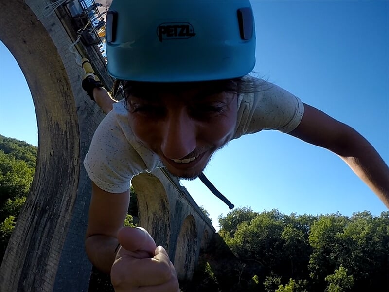 Un gros plan d'une personne souriante avec un casque bleu, suspendue par un élastique au-dessus d'un pont et d'une forêt.