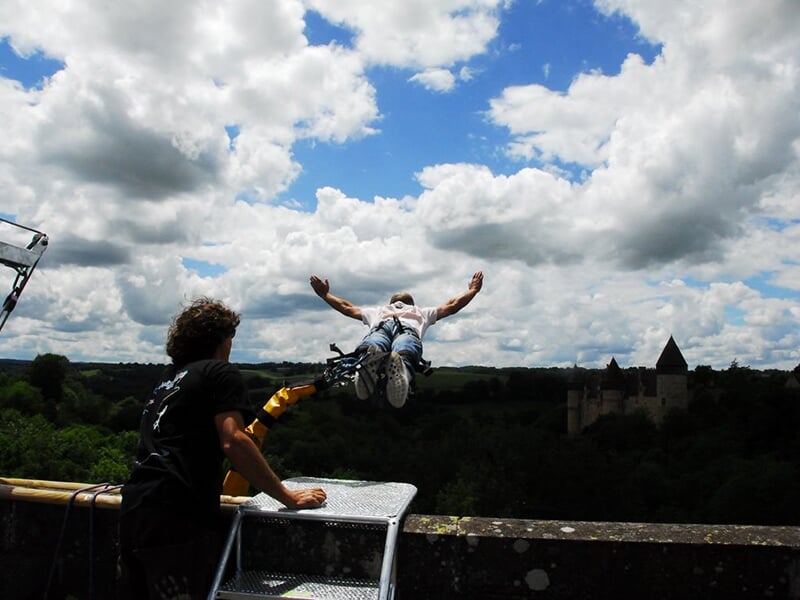 Un homme effectuant un saut à l'élastique, bras ouverts, avec un château en arrière-plan sous un ciel nuageux.