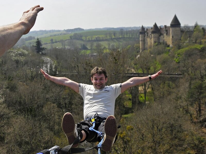 Un homme souriant suspendu lors d'un saut à l'élastique près de Clermont-Ferrand, avec le château de Culan et des collines verdoyantes en arrière-plan.