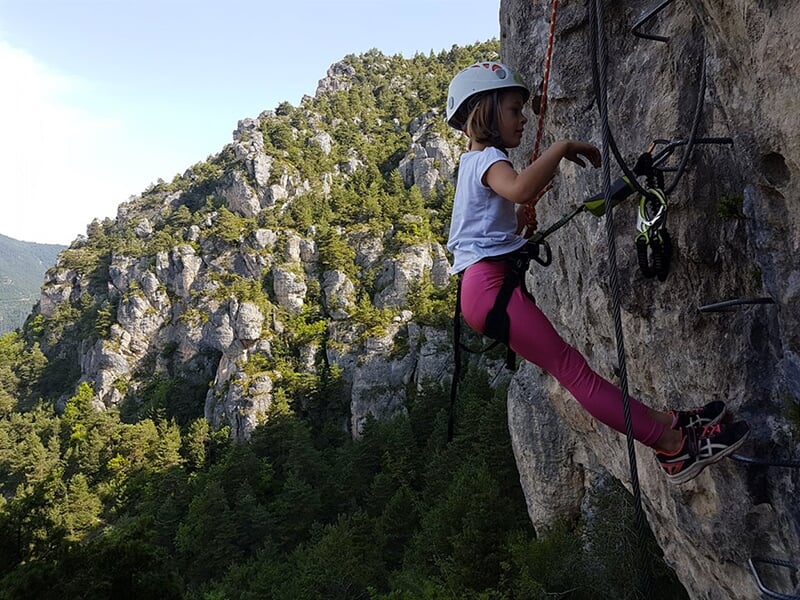 Jeune fille en tenue d'escalade sur une via ferrata, utilisant des barreaux métalliques fixés sur une paroi rocheuse, entourée d'une forêt dense et de montagnes.