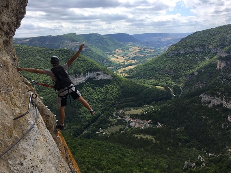 Homme en équilibre sur une via ferrata, tenant un câble, avec une vue panoramique sur une vallée verdoyante et des collines en arrière-plan.