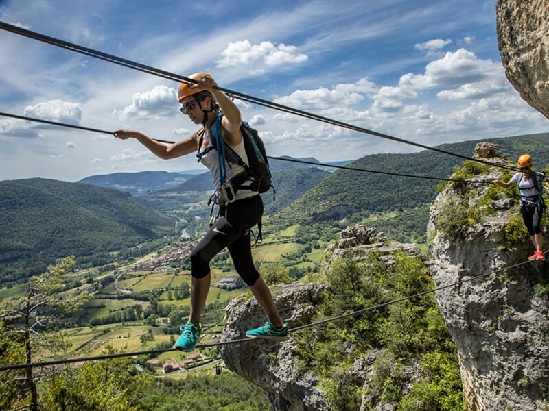 Femme traversant un pont de câbles au-dessus d'un paysage montagneux, avec un village visible au loin dans une vallée verdoyante.