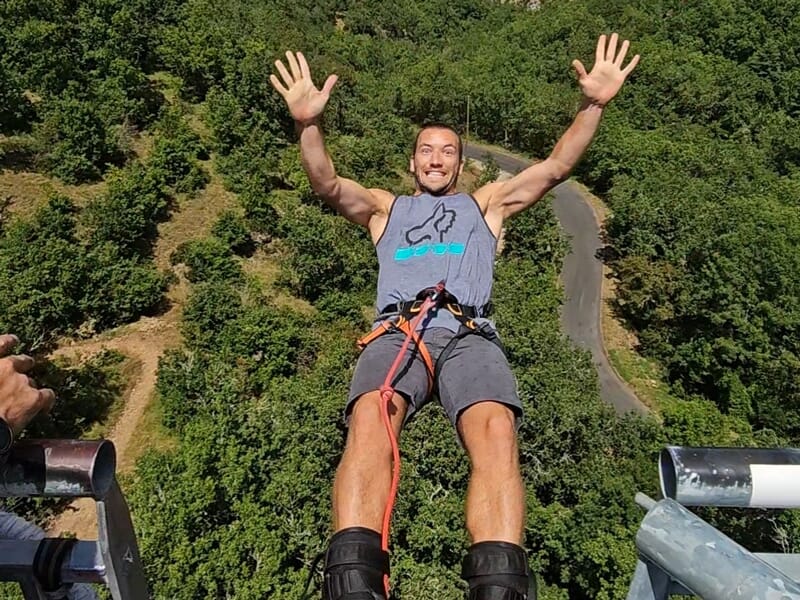 Homme souriant en tenue sportive prêt à sauter à l'élastique avec une forêt dense en arrière-plan.