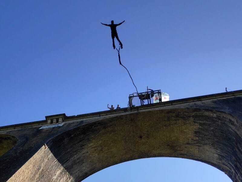 Silhouette d'une personne en plein saut à l'élastique depuis un pont en pierre sous un ciel bleu clair.
