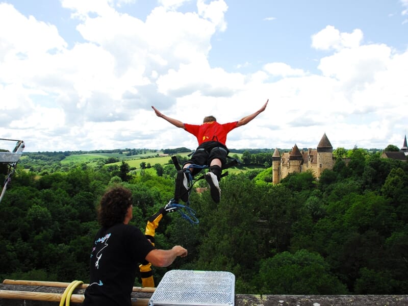 Personne en plein saut à l'élastique depuis un viaduc avec un château et une forêt en arrière-plan sous un ciel bleu.