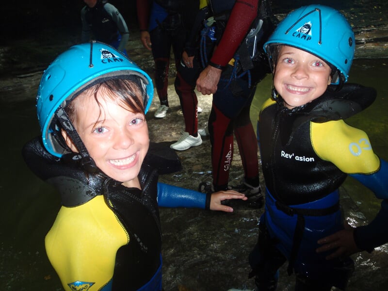 Deux enfants souriants en combinaison et casque bleu prêts pour une activité de canyoning en rivière.