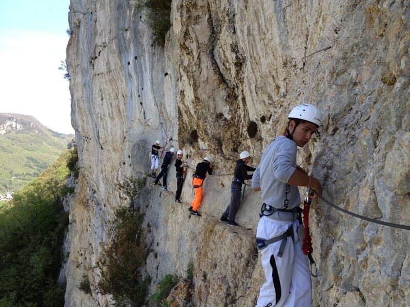 Groupe de personnes équipées de casques et harnais progressant sur une via ferrata accrochée à une falaise rocheuse.