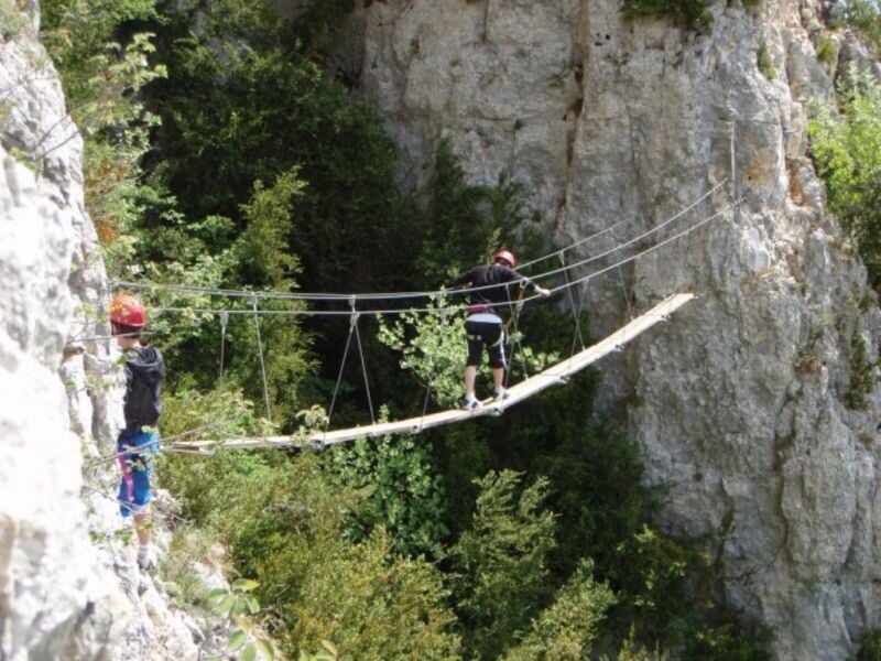 Personne traversant un pont suspendu en bois entre deux falaises dans un cadre naturel verdoyant.