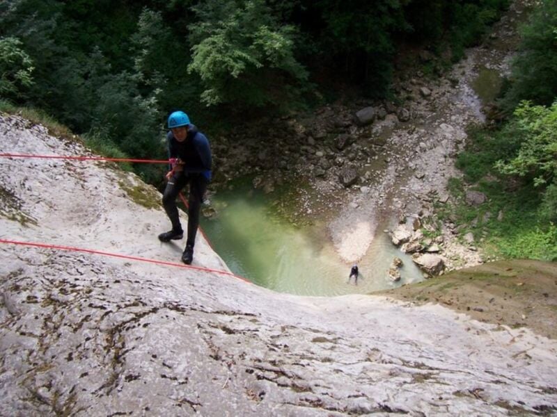 Personne en tenue de canyoning descendant en rappel une paroi rocheuse au-dessus d'un bassin d'eau.