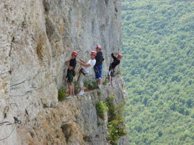 Quatre personnes équipées de casques et harnais marchant sur une via ferrata le long d'une falaise avec vue sur la vallée.