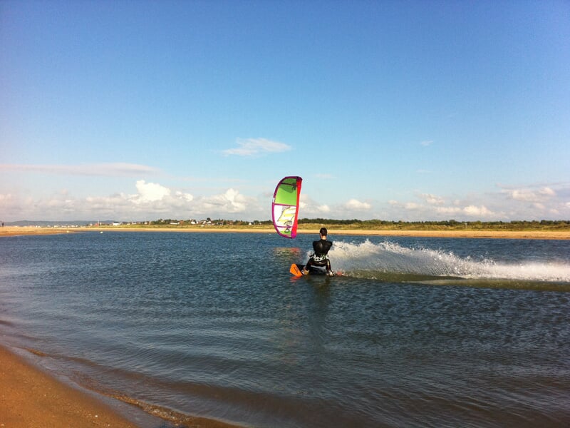 Un kitesurfeur glisse sur l'eau près du rivage sous un ciel bleu clair avec un cerf-volant coloré.
