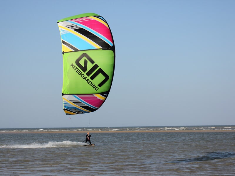 Un kitesurfeur avance sur l'eau avec un grand cerf-volant vert, rose et jaune sous un ciel bleu.