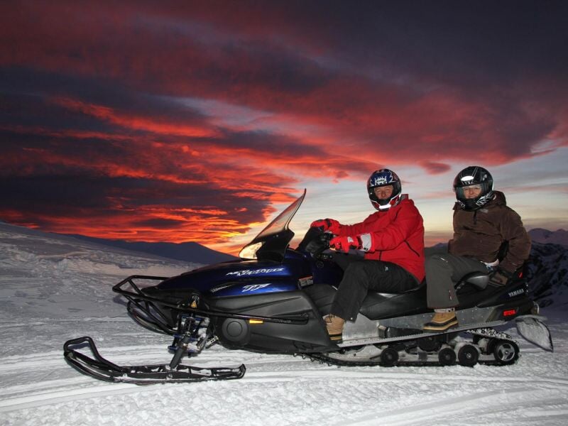 Deux personnes en tenue d'hiver sur une motoneige noire dans la neige au coucher du soleil rougeoyant.