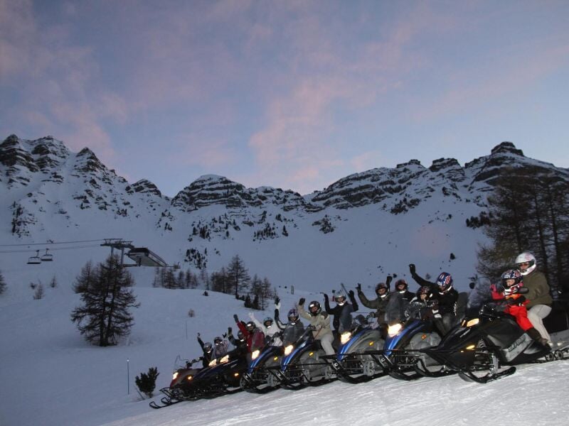 Groupe de motoneiges alignées sur la neige avec des conducteurs levant les bras devant des montagnes enneigées.