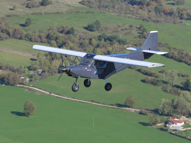 Avion léger gris en vol au-dessus d'un paysage rural verdoyant, avec des champs et des arbres en arrière-plan.