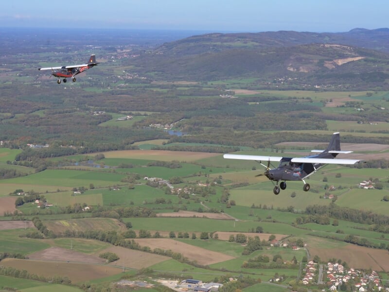 Deux avions légers en formation volant au-dessus d'une vaste campagne verte avec des collines, des champs et des habitations dispersées.